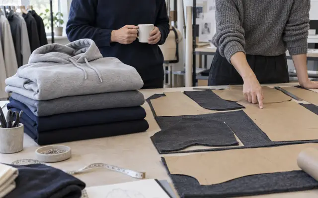 Ready-made hoodies stacked beside paper patterns and cut fleece panels on a workshop cutting table