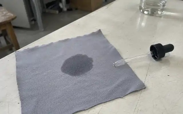 Damp patch spreading on grey jersey beside a glass dropper