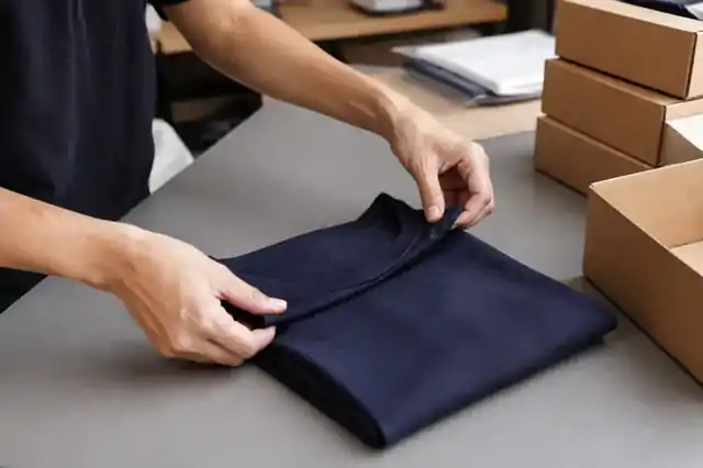 Navy T-shirt being folded at a packing table beside plain cartons